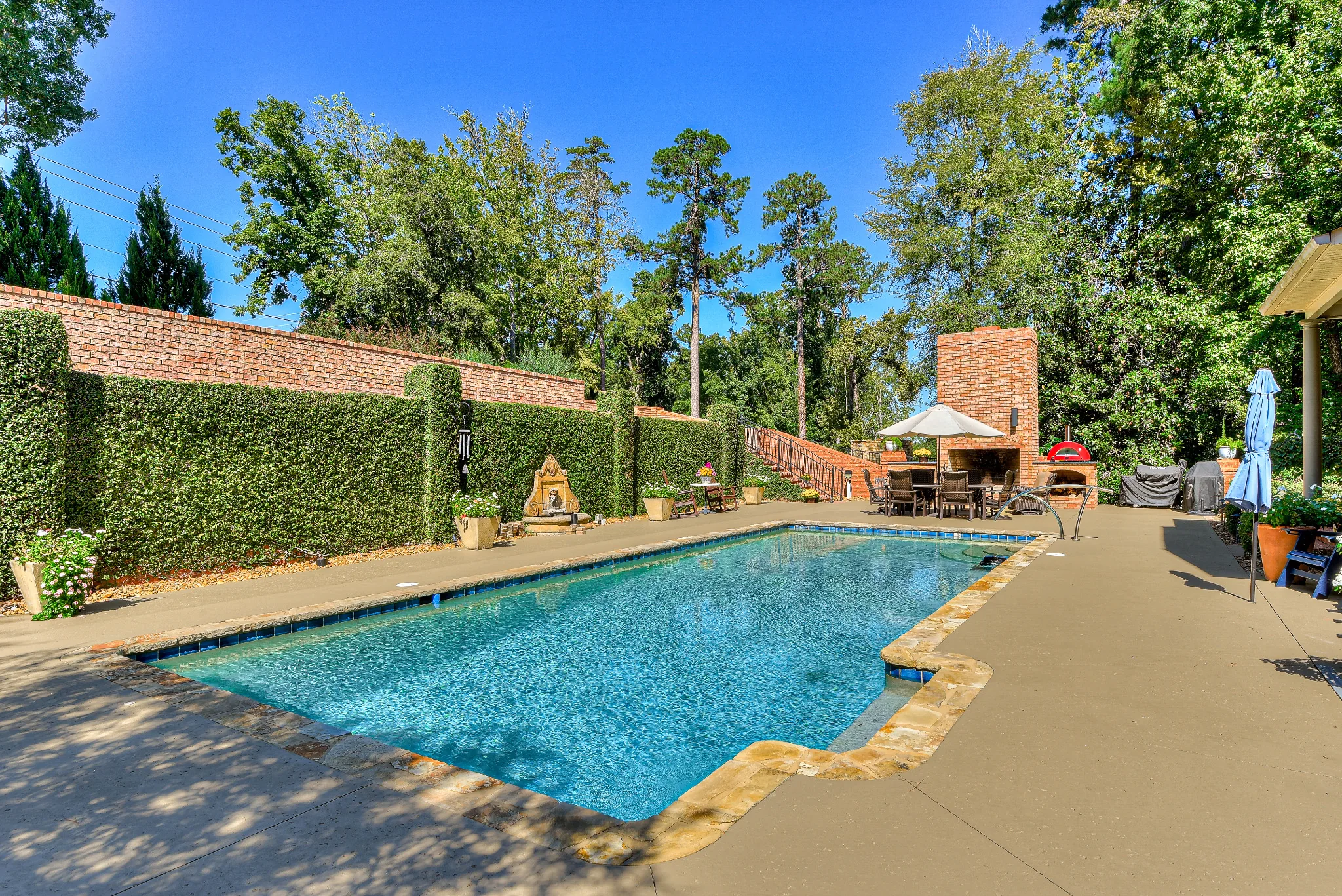 Pool terrace bordered by greenery with an outdoor fireplace and dining area
